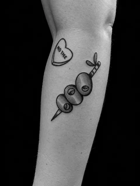 Tatuaje tradicional de aceitunas en un palillo de cóctel y un corazón con la frase «No idea» en un antebrazo