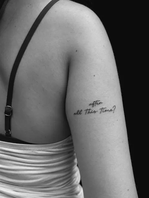 Tatuaje de letra cursiva con la frase «after all this time?» en la parte superior del brazo
