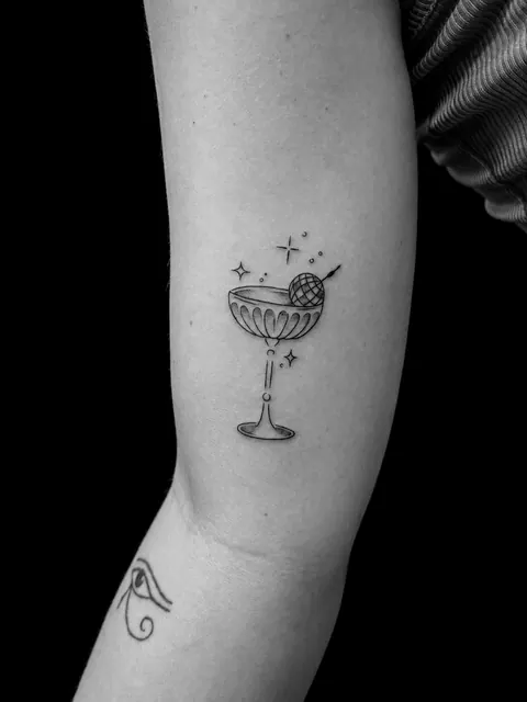 Tatuaje fine line de una copa de cóctel con destellos en un antebrazo