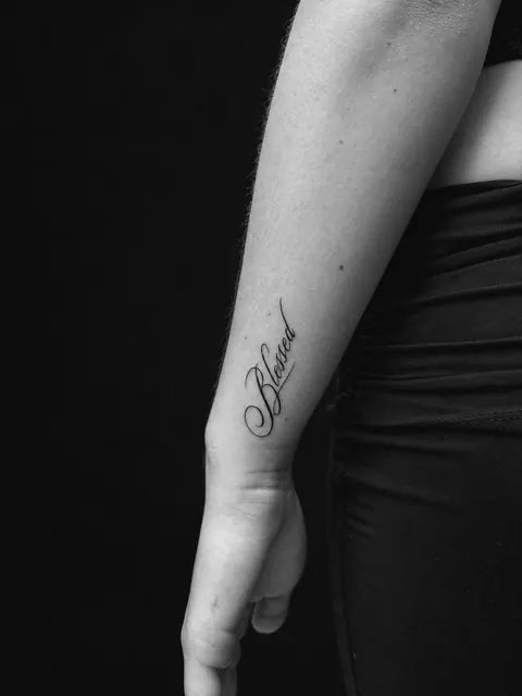Tatuaje de letra cursiva con la palabra «Blessed» en un antebrazo