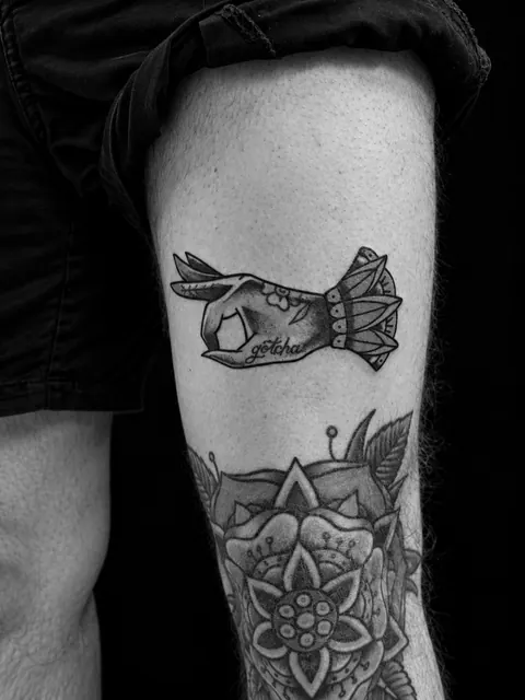 Tatuaje tradicional de una mano haciendo el gesto de OK con la palabra «gotcha» en una espinilla