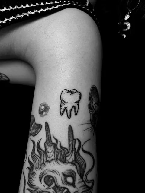 Tatuaje de un diente al estilo grabado en la parte superior del brazo, entre otros tatuajes más antiguos de otros artistas