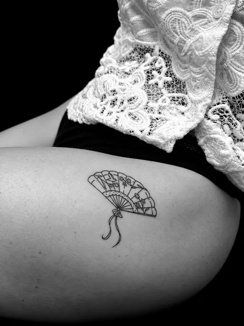 Tatuaje fine line de un abanico español con flores y una borla en un muslo