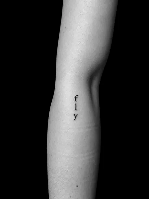 Pequeño tatuaje vertical con la palabra «fly» en letra con serifa en la parte interior del brazo