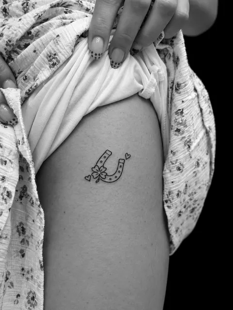 Tatuaje de línea fina de una herradura con un trébol y dos corazones pequeños en un muslo