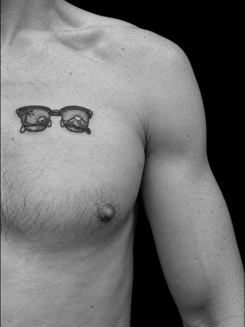 Tatuaje de unas gafas de sol en el pecho, con una playa y una palmera reflejadas en los cristales