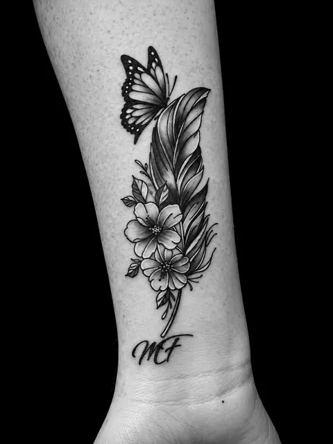 Tatuaje ilustrativo de una pluma con flores, una mariposa y lettering, en negro y gris, en una espinilla