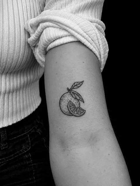 Tatuaje pequeño de un cítrico con hoja y una rodaja, sombreado con puntos finos, en la parte superior de un brazo