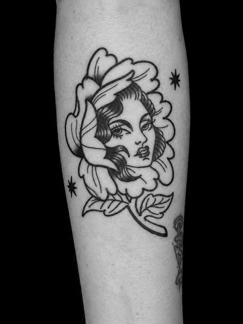 Tatuaje tradicional del rostro de una mujer dentro de una flor, con dos estrellas pequeñas, en un antebrazo