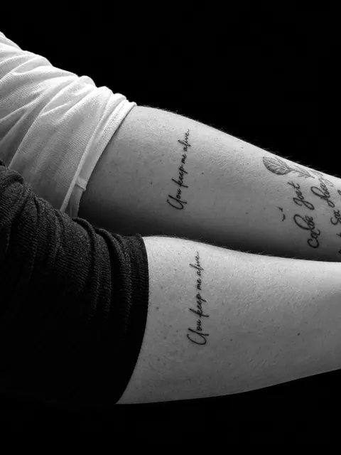 Dos frases iguales en cursiva vertical «You keep me alive» en dos antebrazos