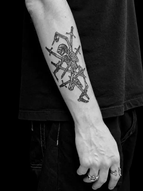 Tatuaje de estilo grabado con un esqueleto atravesado por espadas en un antebrazo
