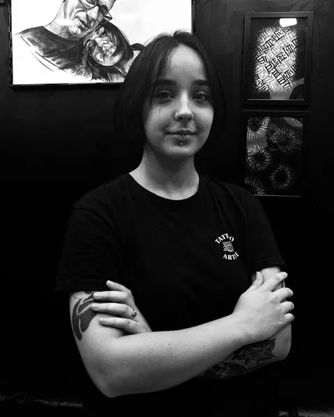 Margo en el estudio, con los brazos cruzados, dibujos de tatuaje enmarcados en la pared detrás de ella